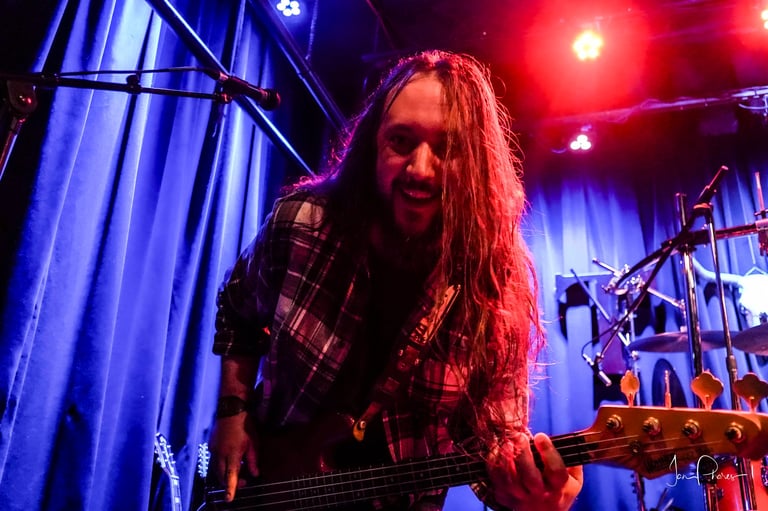 Musician with long hair playing bass guitar on stage under blue and red stage lights during a live concert performance