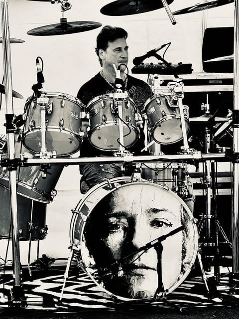 Black and white photograph of a drummer performing at a drum kit with a portrait image printed on the bass drum