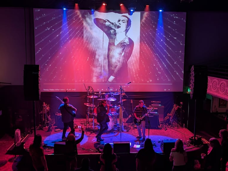 Live band performing on stage with pink and blue lighting, large projection screen displaying artist image above, audience in foreground