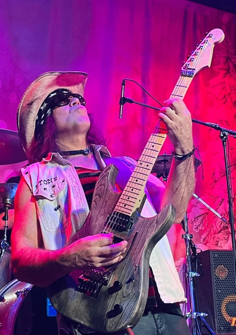 Musician wearing cowboy hat and sunglasses plays electric guitar on stage under vibrant pink and purple lighting