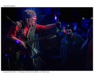 Guitarist performing on stage wearing red shirt and patterned headwrap, playing to audience in dimly lit venue with blue lighting