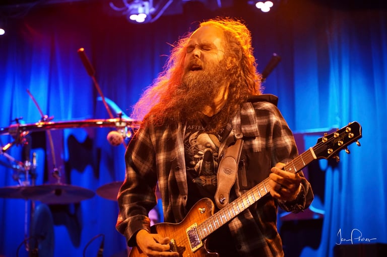 Bearded musician with long red hair performing with electric guitar on stage under blue stage lighting