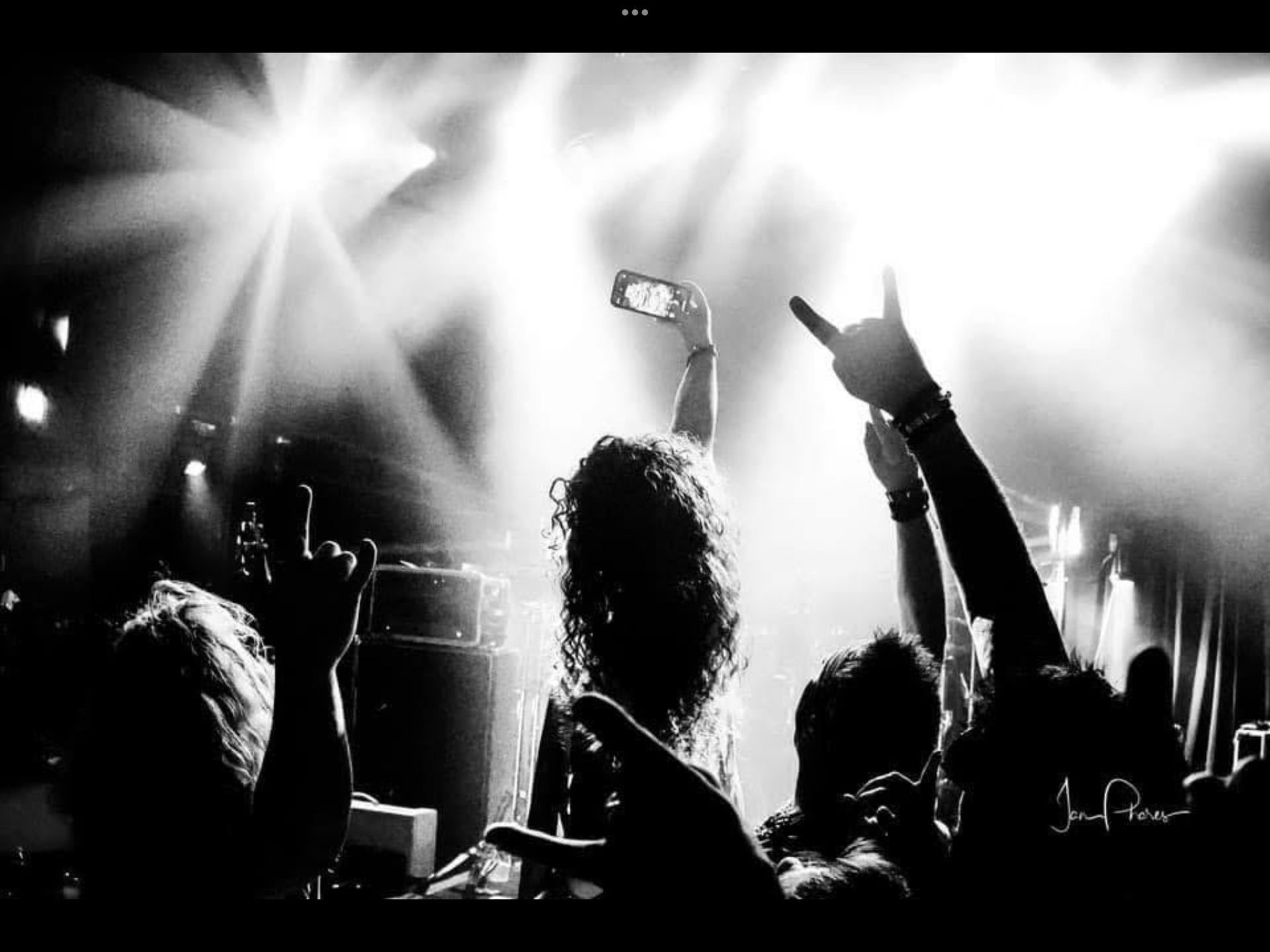 Black and white concert photo showing silhouetted crowd with raised arms and phones under dramatic stage lighting