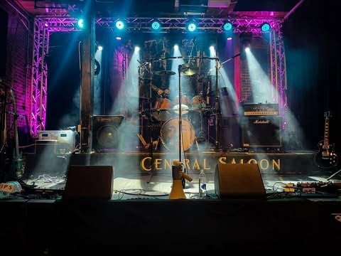 Live concert stage with dramatic purple and blue lighting, band performing with drum kit, scaffolding structure, and Central Saloon signage visible
