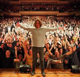 Person standing on stage with arms outstretched facing a large crowded auditorium with audience members raising hands and cheering