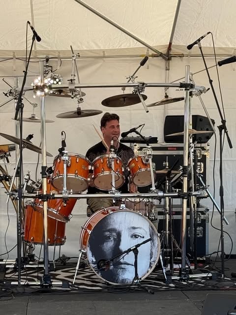 Drummer performing at an outdoor concert under a white tent canopy with professional stage lighting and equipment
