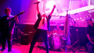 Live band performance on stage with purple and pink lighting, featuring musicians and audience members with arms raised