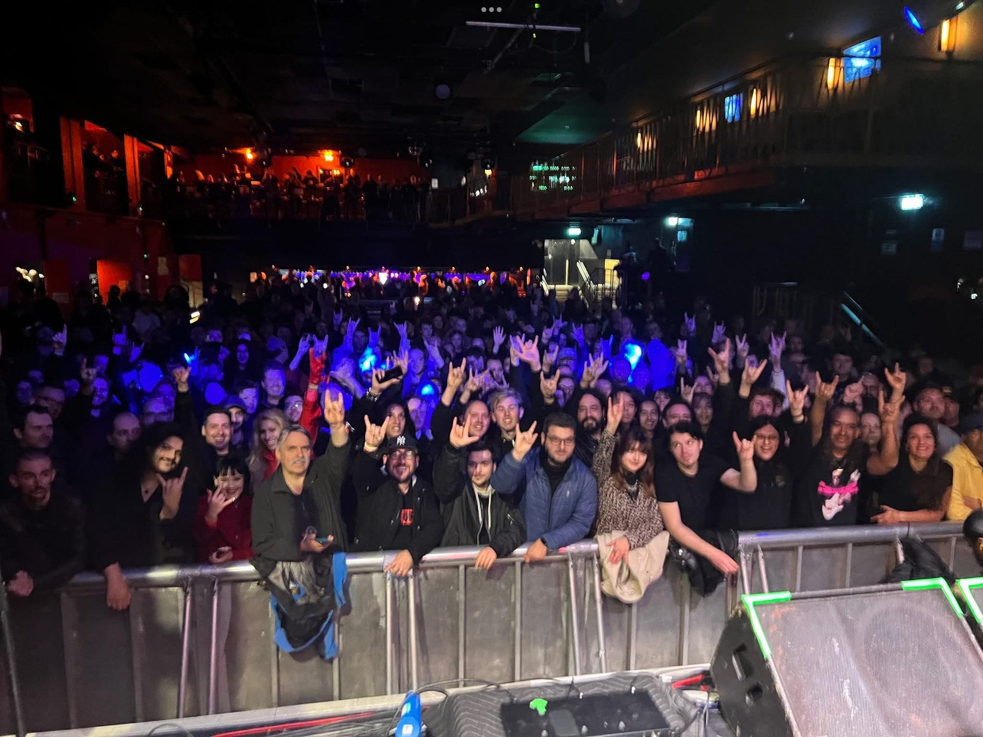 Large crowd of people at a concert venue with blue and red stage lighting and barriers in the foreground