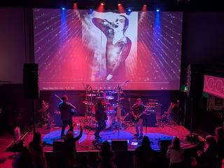 Live band performing on stage with red and blue lighting, large video screen displaying performer above, and audience in foreground