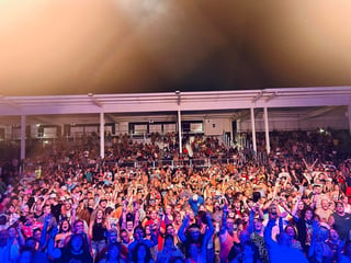 Large crowd at an indoor concert or event venue with colorful stage lighting and white structural supports overhead