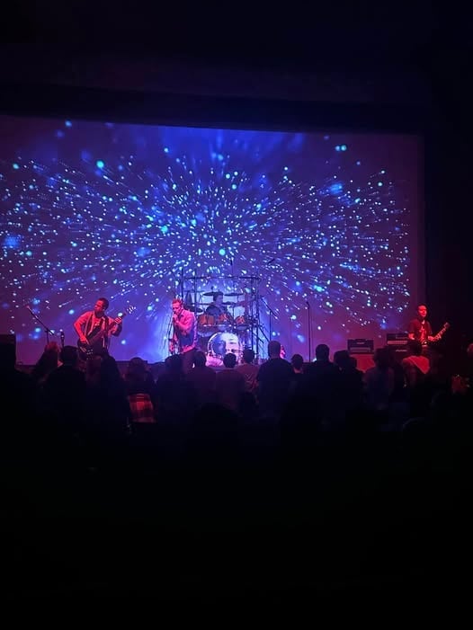Live concert performance on stage with band members silhouetted against blue and purple lights with animated particle effects projection
