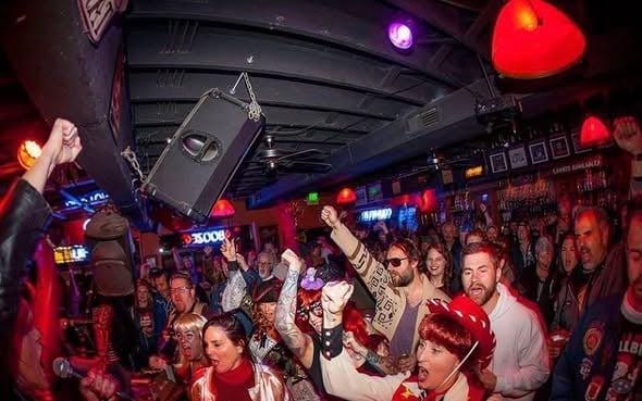 Crowded nightclub scene with people dancing under red and blue lights, suspended speaker overhead, and DJ booth visible